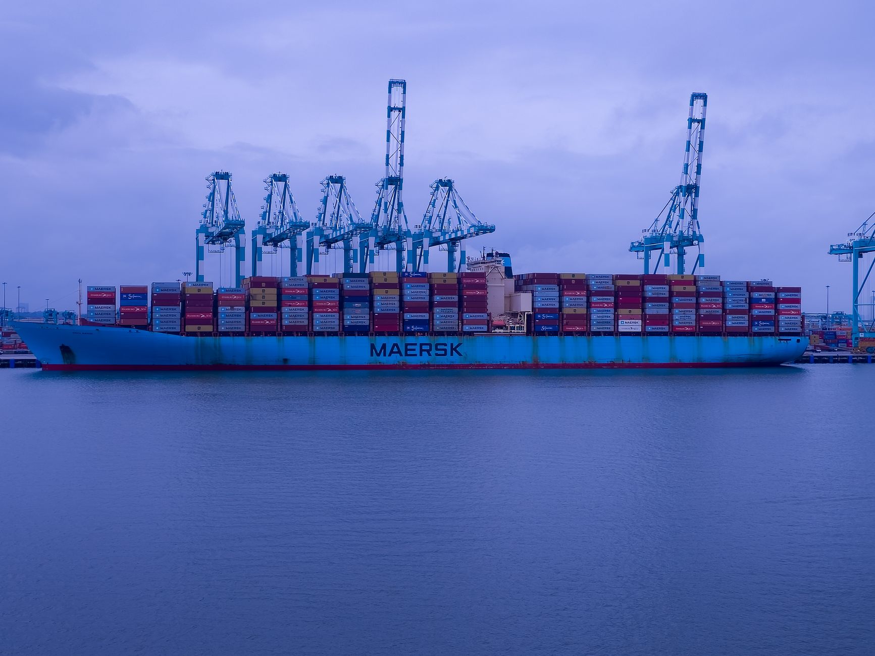 A Maersk cargo ship loaded with containers at an industrial port under a cloudy sky.