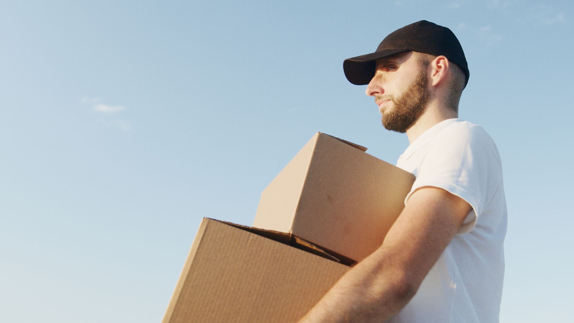 Deliveryman with beard and black hat holds boxes outdoors in daylight.