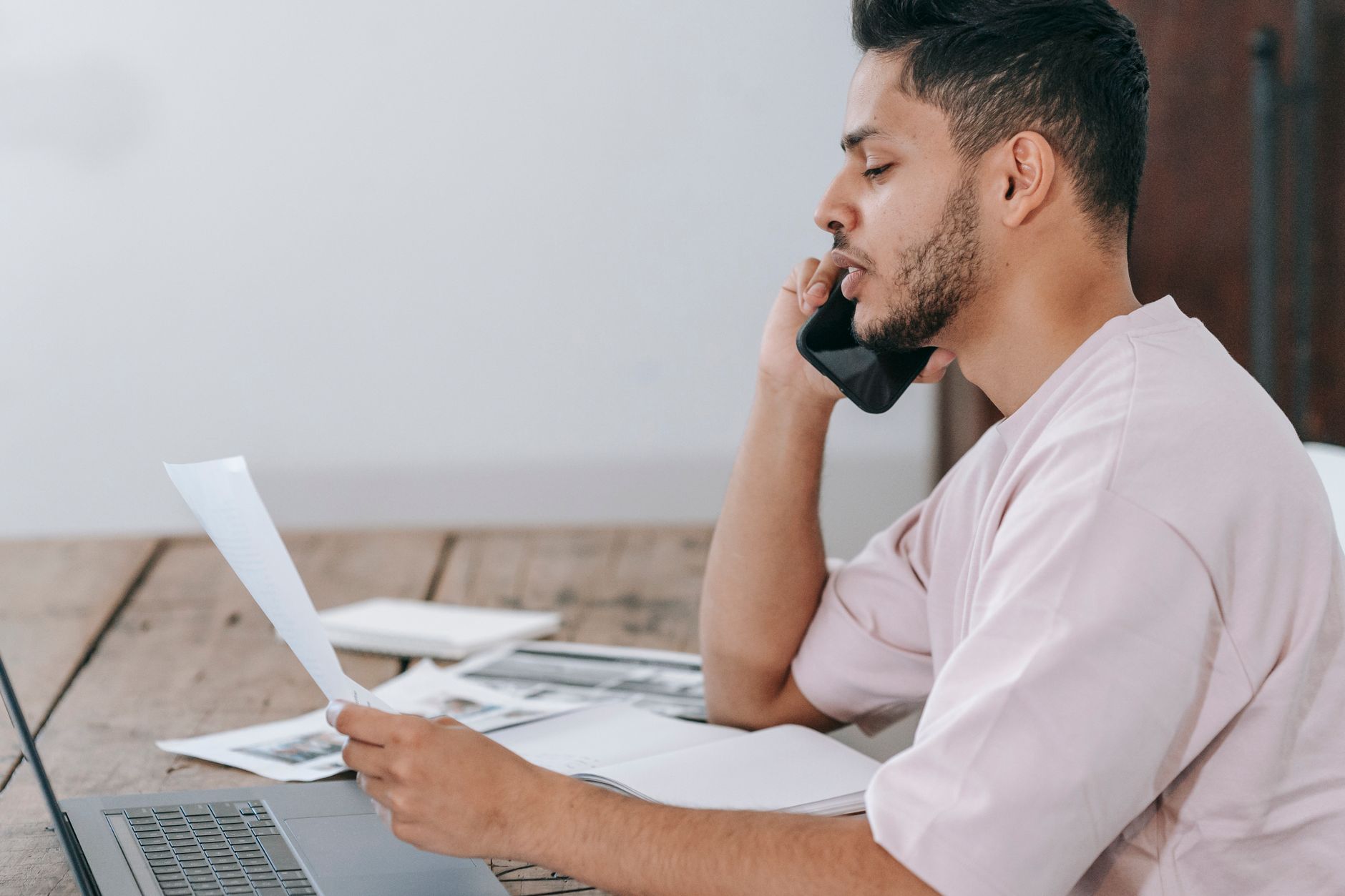 Side view of young Hispanic male remote employee discussing documents during phone call while working online with laptop and papers in home 