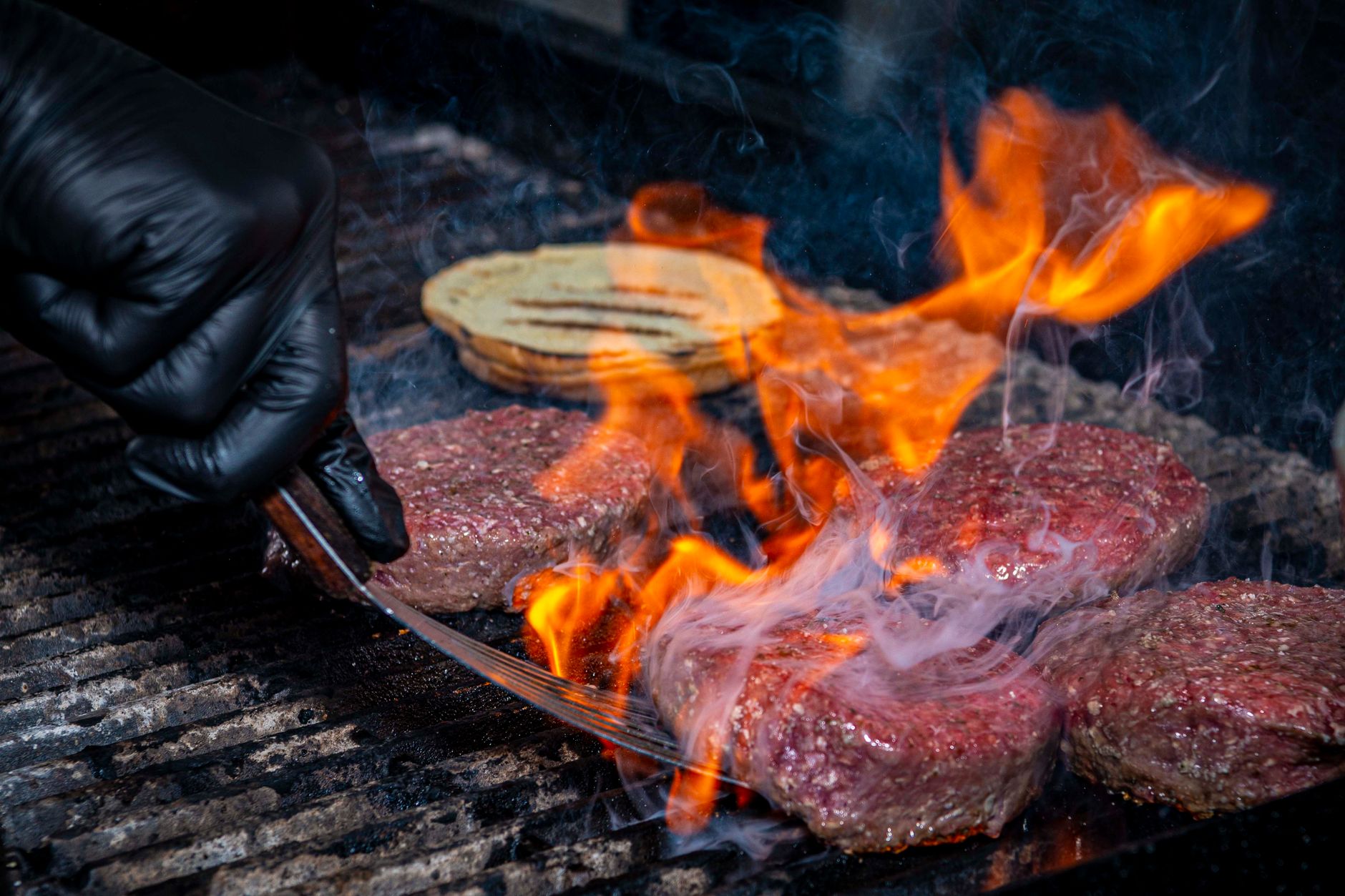Black-gloved hand grilling burgers with flames on a barbecue grill.