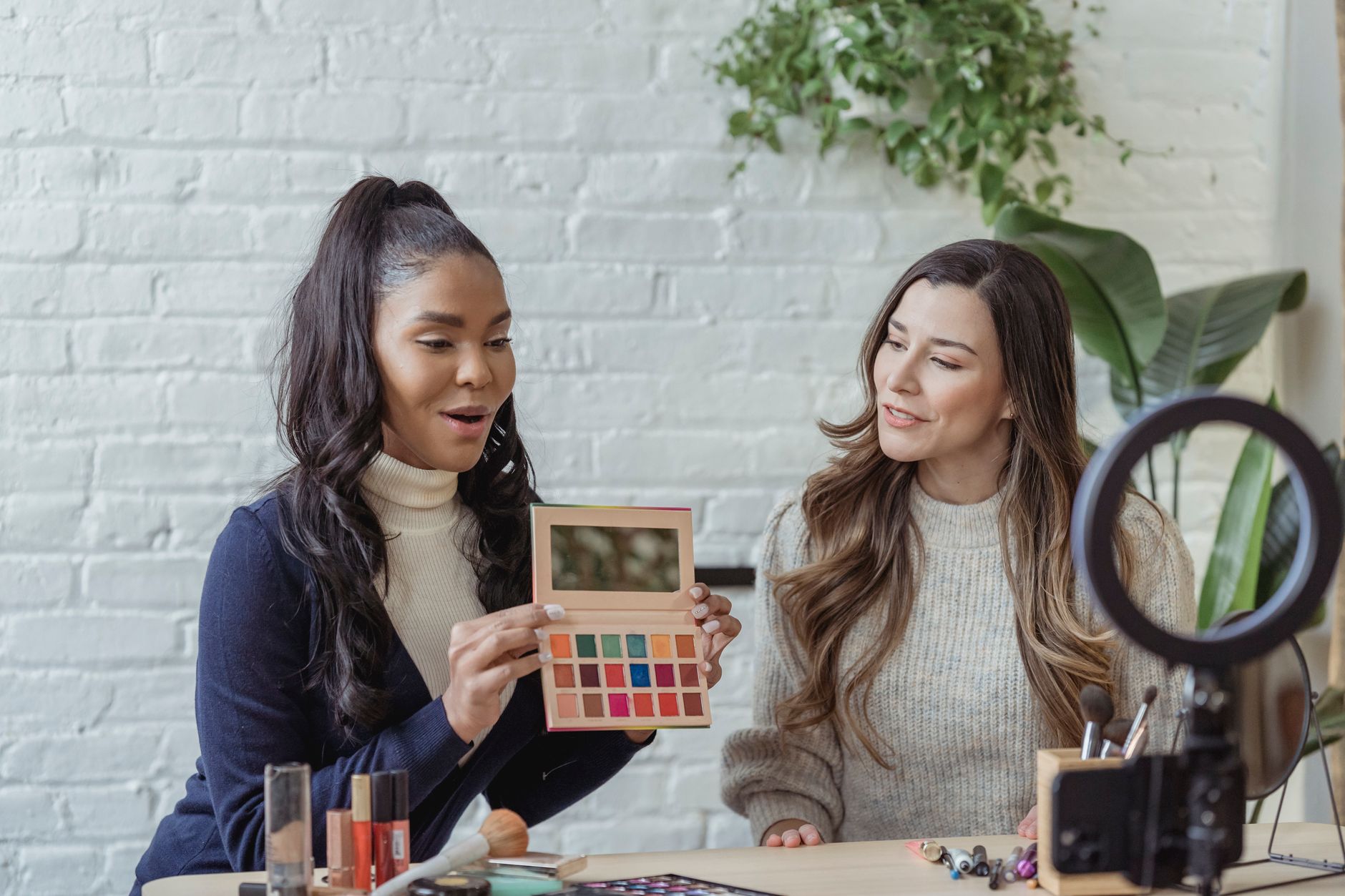 Young African American female blogger demonstrating palette of colorful eyeshadow while recording vlog review on smartphone placed on tripod
