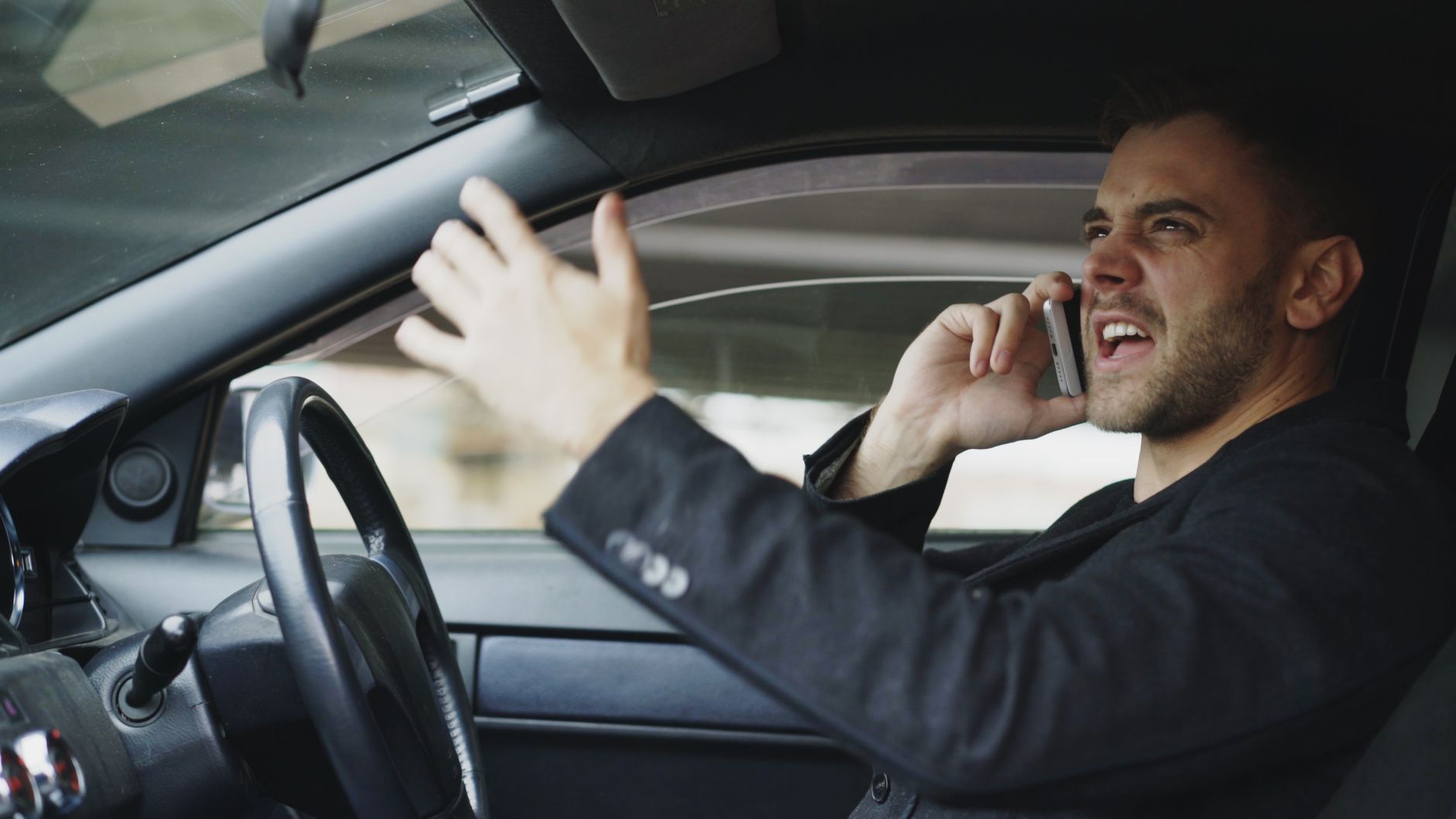 Stressed man gesturing while on phone in car, expressing frustration.