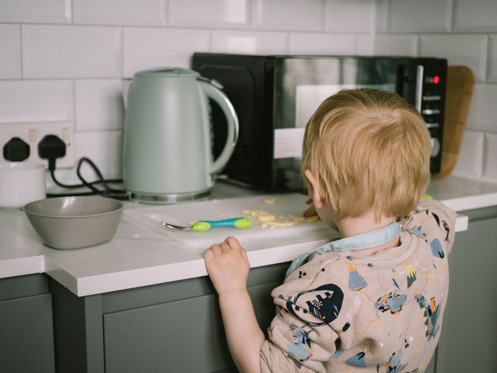 A toddler reaching for food on the kitchen counter next to a kettle and microwave.