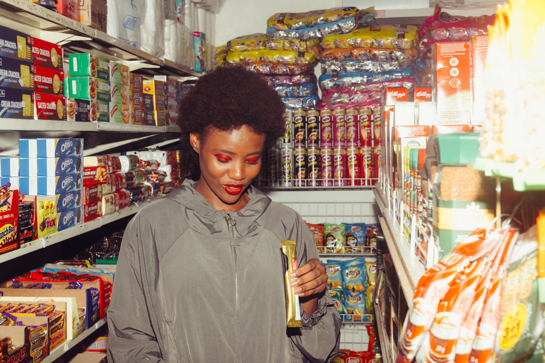 Adult black female buyer in casual outerwear with bright makeup holding chocolate product while standing in supermarket and choosing goods f