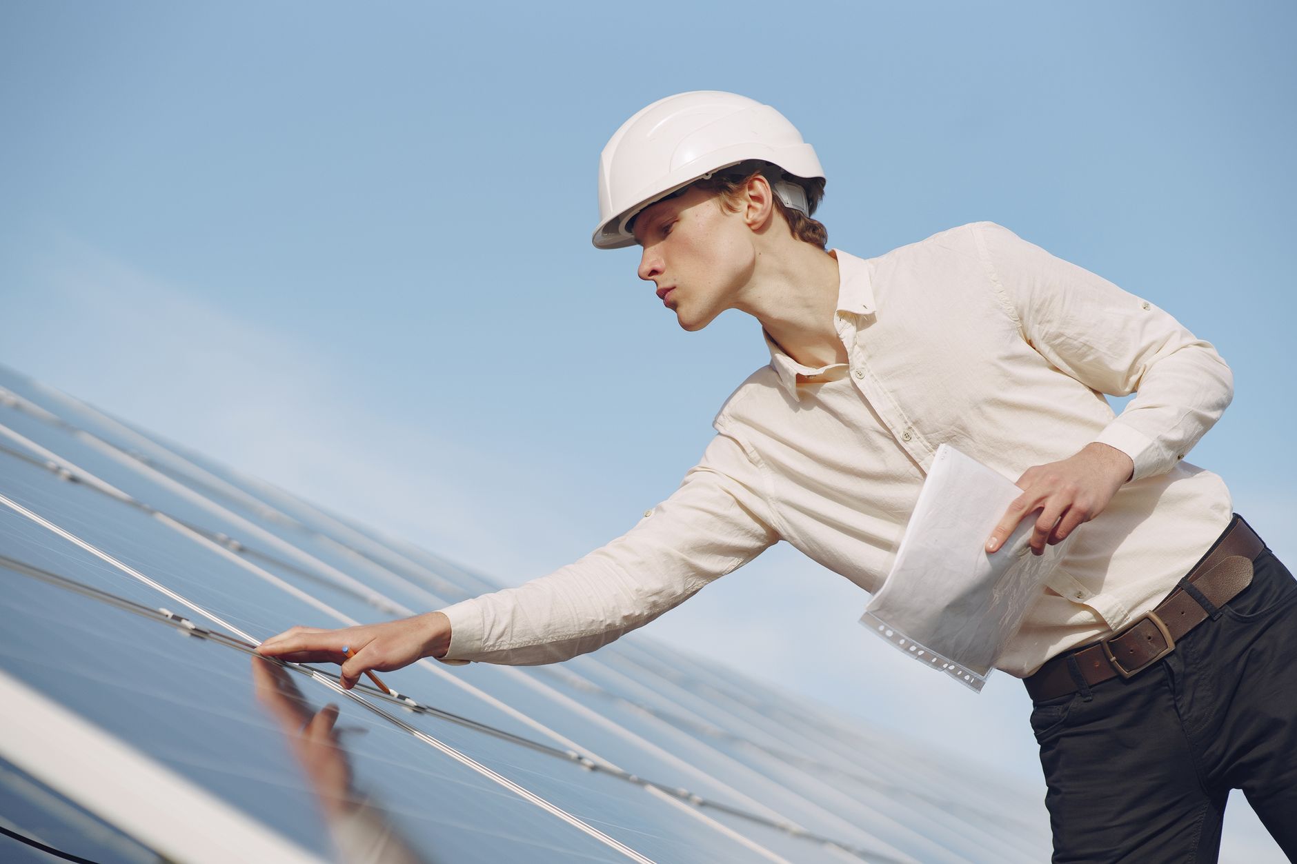 Low angle of young male inspector with documents in formal wear and hardhat standing against solar panels and checking details of constructi