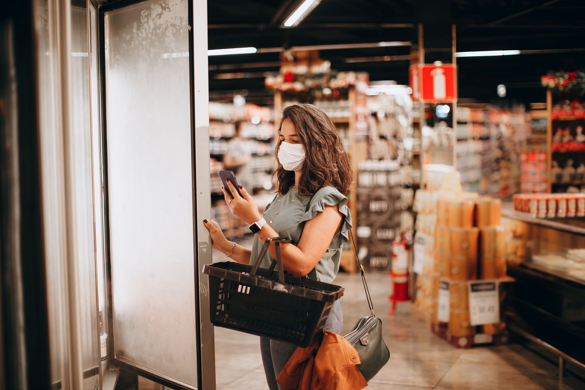 Woman wearing mask shopping with a basket and smartphone in a supermarket.