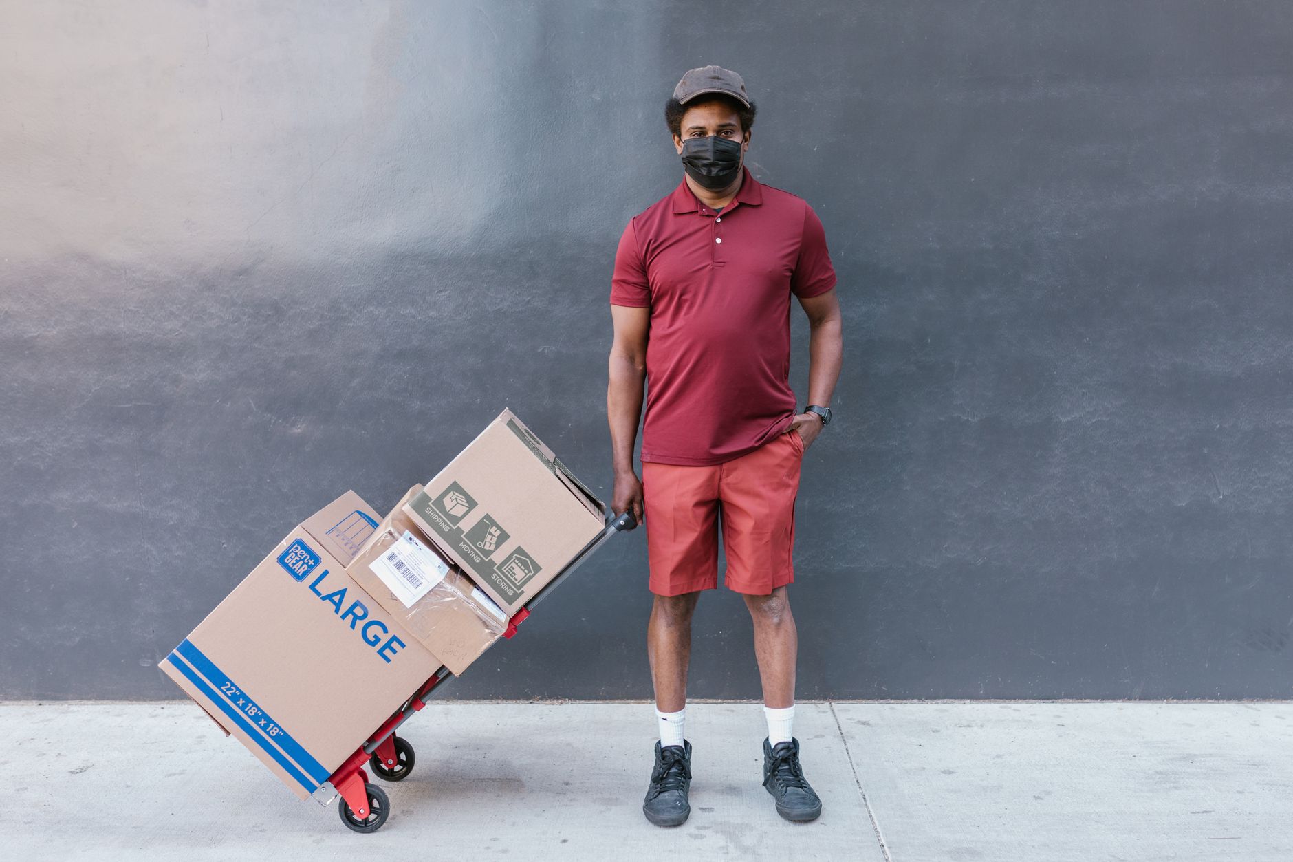 Man delivering boxes via hand truck, standing against a gray wall outdoors.